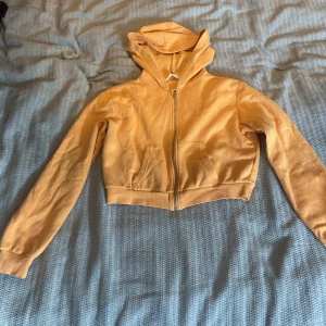 Gul croppad hoodie från H&M - Snygg gul croppad hoodie från H&M med dragkedja framtill och huva. Tröjan har långa ärmar, ribbade muddar och två fickor framtill. Perfekt för dig som gillar en avslappnad och trendig stil.