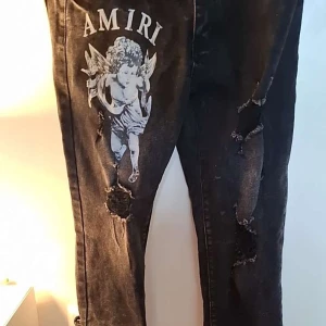 Svarta Amiri jeans med ängeltryck - Säljer ett par svarta jeans från Amiri med stort vitt ängeltryck på ena benet och flera slitningar och hål för en edgy look. Jeansen har klassisk femficksdesign och Amiri-logga på bakfickan. Perfekta för dig som gillar streetstyle och detaljer.