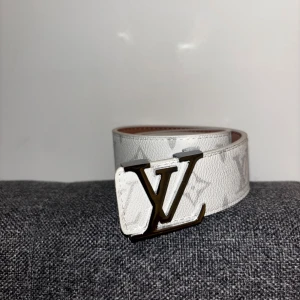 Vit Louis Vuitton monogram bälte - Snyggt vitt bälte från Louis Vuitton med klassiskt monogrammönster och stor LV-logga i metall som spänne. Bältet är brett och gjort i präglat skinn med diskreta grå detaljer. Perfekt accessoar för att lyfta din outfit.