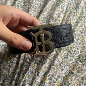 Burberry svart skärp med TB-spänne - Snyggt svart skärp från Burberry med stort metallfärgat TB-spänne framtill. Utsidan har präglat mönster och insidan har klassiskt rutigt Burberry-tyg i beige, rött och svart. Perfekt accessoar för att lyfta din outfit.