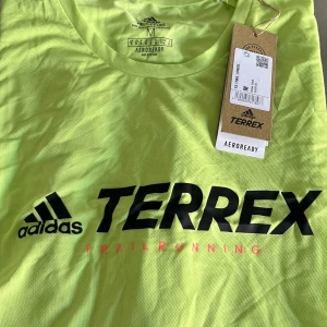 Adidas Terrex långärmad träningströja - Neongul långärmad träningströja från Adidas Terrex med svart logga och texten 'Trail Running' framtill. Tillverkad i lätt och ventilerande polyester, perfekt för löpning eller annan träning. Regular fit och rund halsringning.
