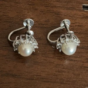 Vintage örhängen i 14K vitguld med pärlor - Snygga vintage örhängen i 14K vitguld med skruvstängning. Varje örhänge har en rund vit pärla i mitten, omgiven av små gnistrande stenar i en elegant korgformad infattning. Perfekt för dig som gillar klassisk och tidlös stil.