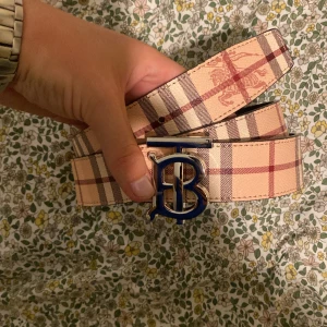 Burberry bälte  - Snyggt bälte från Burberry i klassiskt rutigt mönster med beige, bruna och röda toner. Bältet har ett stort metallspänne med blått B, vilket ger en lyxig och trendig känsla. Perfekt accessoar för att lyfta din outfit.