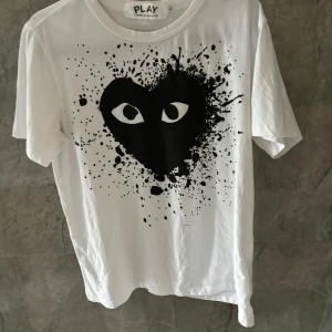 Vit ami paris t-shirt - Säljer en vit t-shirt från Comme des Garçons Play med det ikoniska svarta hjärtat med ögon och coolt splattermönster på bröstet. Klassisk passform och rund halsringning. Perfekt statement-plagg för dig som gillar streetstyle.
