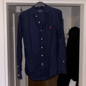 Blå linneskjorta Polo Ralph Lauren S - Snygg marinblå skjorta från Polo Ralph Lauren i slim fit-modell. Skjortan är tillverkad i linne och har vita knappar samt det klassiska röda broderade logot på bröstet. Perfekt för dig som gillar stilrena och tidlösa plagg. 300