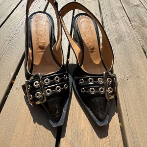 Svarta pumps med spänne från GANNI - Snygga svarta pumps från GANNI med spetsig tå, breda remmar med stora silverspännen och metallöljetter. Skorna har låg klack och är tillverkade i glansigt skinn. Perfekta för dig som gillar statement-detaljer och edgy stil. Säljs inte längre och högefterfrågan därav priset. Nypris 3595kr