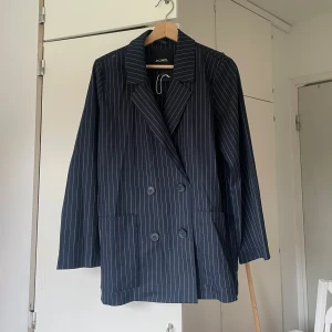 Randig kostym från Monki, mörkblå - Snygg tvådelad kostym från Monki i mörkblått med vita smala ränder. Kavajen är dubbelknäppt med bred krage och stora fickor, byxorna har hög midja och raka ben. Perfekt för dig som vill sticka ut med en klassisk men trendig look.