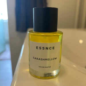 ESSNCE Carashmellow Eau de Parfum - Parfymflaska med gul vätska från ESSNCE, doften Carashmellow. Flaskan är cylindrisk i genomskinligt glas med svart rund kork och vit etikett med svart text. Modern och stilren design som sticker ut i badrumshyllan.