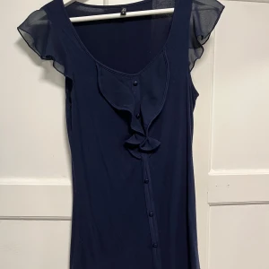 Mörkblå topp med volang från Ann Christine - Mörkblå topp från Ann Christine med transparenta volangärmar och dekorativa knappar framtill. Toppen har en snygg volangdetalj längs urringningen och är tillverkad i ett mjukt, lätt material som känns bekvämt mot huden.