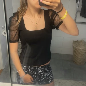 Svart topp med puffiga meshärmar - Snygg svart topp med fyrkantig urringning och korta puffärmar i transparent mesh med små prickar. Kroppsdelen är i mjukt tyg och toppen är croppad. Perfekt för dig som vill ha en trendig och stilren look.