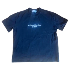 Maison Margiela svart t-shirt med tryck - Svart t-shirt från Maison Margiela med vit logotyp på bröstet och coolt tryck med olika språk på ryggen. Klassisk passform, rund hals och korta ärmar. Perfekt statement-plagg för dig som gillar streetstyle och designer.