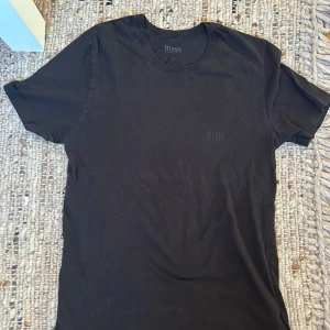 Svart Hugo Boss t-shirt - En stilren svart t-shirt från Boss med diskret broderad logga på bröstet. Klassisk rund halsringning och korta ärmar. Tillverkad i mjuk bomull som känns skön mot huden. Perfekt för en clean och enkel look.