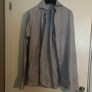 Ljusgrå slim fit skjorta från Eton - Snygg ljusgrå skjorta från Eton i slim fit-modell. Skjortan har långa ärmar, klassisk krage och knappar framtill. Insidan av kragen och knappslån har ett rutigt blåvitt mönster som ger en schysst detalj. Tillverkad i bomull för en skön känsla.