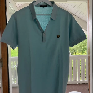 Ljusblå piké från Lyle & Scott - Snygg ljusblå pikétröja från Lyle & Scott med klassisk krage och knappslå. Ikonisk gul logga på bröstet i storlek L men passar M. Perfekt för dig som gillar stilren och enkel design. Skön passform och mjukt material.
