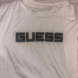 Vit t-shirt från Guess med logga - Vit t-shirt från Guess med stor svart logga framtill. Klassisk rund halsringning och korta ärmar. Tillverkad i mjuk bomull som känns skön mot huden. Enkel och clean stil som passar perfekt till jeans eller shorts.