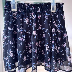 Blommig svart volangkjol - Supersöt kort kjol med svart botten och blommigt mönster i rosa och blått. Kjolen har volanger och resår i midjan för en bekväm passform. 