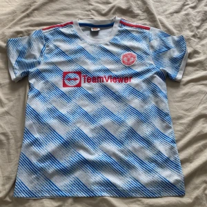 Manchester United fotbollströja  - Manchester United bortatröja med Ronaldo 7 på ryggen. Tröjan är vit med blått diagonalt mönster, röda detaljer och klubbmärke på bröstet. Adidas-logga och TeamViewer sponsortryck. Klassisk rund hals och korta ärmar. Perfekt för fans av Ronaldo och United. Storlek 30