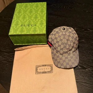 Gucci keps med GG-mönster, storlek M - Fast pris!!! Exklusiv keps från Gucci i beige med klassiskt GG-monogram över hela kepsen. Kepsen har en böjd skärm, justerbar rem bak och ikoniska röd-blå ränder på sidan. Tillverkad i bomullscanvas och levereras med originalbox och dustbag.