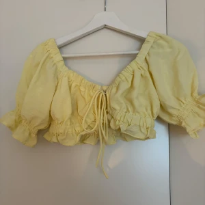 Gul croppad blus från Gina Tricot - Supersöt ljusgul croppad blus från Gina Tricot med puffiga korta ärmar, volangkant och snörning framtill. Perfekt för varma dagar med sin luftiga känsla och trendiga offshoulder-look. Materialet är mjukt och lätt, vilket gör den extra bekväm.