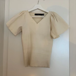 Beige topp med puffärm från Vero Moda - Superfin beige topp från Vero Moda med v-ringning och korta puffärmar. Toppen har en mjuk, stickad känsla och är figurnära i modellen. Perfekt för dig som gillar en enkel men ändå trendig look med volym på axlarna. Lite fläckig som säkert går bort i tvätten - därav priset.