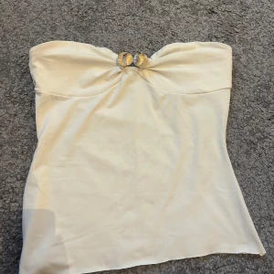Vit tubtopp med gulddetalj - Snygg vit tubtopp med en elegant guldfärgad detalj framtill som ger en lyxig känsla. Toppen är axelbandslös och har en enkel, stilren design som passar perfekt till sommarens outfits.