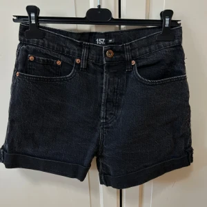 Svarta jeansshorts från 157 - Svarta jeansshorts från 157 med klassisk femficksdesign och uppvikta benslut. Shortsen har en mörk tvätt och kopparfärgade nitar vid fickorna. Perfekta för en chill sommarlook och passar till allt. Använda fåtalgånger, som nya