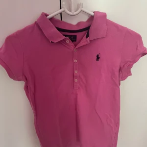 Rosa piké från Polo Ralph Lauren - Snygg rosa pikétröja från Polo Ralph Lauren med klassisk krage, korta ärmar och knappar framtill. Passar mig perfekt och jag är 158cm lång och har storleken xs. Den IKONISKA loggan broderad på bröstet. Perfekt för en sportig och stilren look. Tillverkad i mjuk bomull som är skön att bära hela dagen.