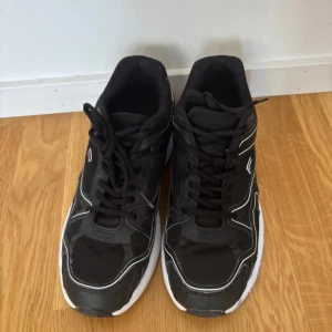 Svarta sneakers med vit sula - Svarta sneakers med vit sula och snygga vita detaljer längs sidorna. Ovandelen är i mesh och syntet, vilket ger en sportig vibe och bra andningsförmåga. Klassisk snörning och rund tå gör dem perfekta för en aktiv vardag.
