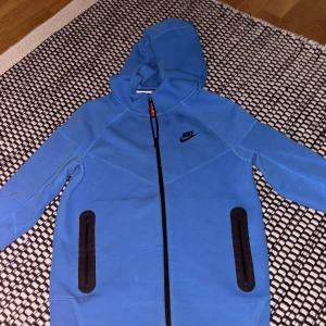 Blå Nike hoodiejacka med dragkedja - Nike hoodiejacka i klarblå färg med svart logga på bröstet. Jackan har hel dragkedja framtill, två svarta fickor med dragkedja och en skön huva. Nike teck i storlek 158-164 vilken motsvara xs i vuxen storlek 