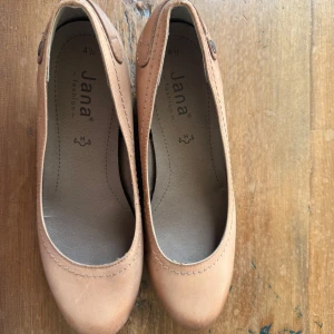 Beige ballerinaskor från Jana - Snygga ballerinaskor från Jana i beige med rund tå och diskret söm längs kanten. Skorna har en enkel och stilren design med liten knappdetalj på sidan. Perfekta för dig som gillar klassisk och clean stil.