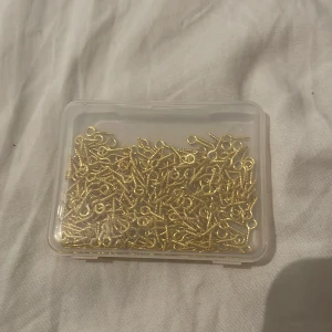 Guldfärgade ögelskruvar till smycken - En ask med massor av små guldfärgade ögelskruvar, perfekta för att göra egna smycken som halsband, armband eller örhängen. Skruvarna är i metall och har en snygg glansig finish. Perfekt för dig som gillar att pyssla och skapa egna accessoarer.