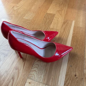Röda pumps med spetsig tå och klack - Snygga röda pumps med glansig finish och spetsig tå. Skorna har hög klack och en klassisk siluett som verkligen sticker ut. Perfekta för dig som vill ha en färgklick till outfiten. Materialet är syntet och insidan är ljusbeige.