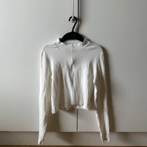 Vit ribbad kofta från Pieces - Säljer en vit ribbad kofta från Pieces med dragkedja framtill. Tröjan har lång ärm, croppad passform och stårkrage. Perfekt att slänga över en topp för en clean och fräsch look. Materialet är mjukt och stretchigt, vilket gör den superbekväm.