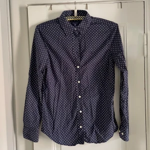 Mörkblå prickig skjorta från Gant - Snygg mörkblå skjorta från Gant med vita prickar och klassisk krage. Den är i fint skick, storlek 36 och något figursydd. Skjortan har långa ärmar, knappar framtill och broderad Gant-logga på bröstet. Tillverkad i mjuk bomull som känns skön mot huden. Perfekt för dig som gillar stilrena mönster och klassisk look💙