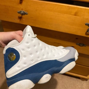 Air Jordan 13 Retro French Blue - Nike Air Jordan 13 Retro med vit ovandel i skinn, blå mockadetaljer och grå tå. Röd Jumpman-logga på plösen och hologramdetalj vid hälen. Klassisk siluett med snörning och rund tå. Perfekta för dig som vill sticka ut med en ikonisk sneaker. Använda 5-10 gånger.  Marketprice 4000-4800 kr