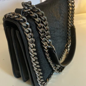 Svart axelväska med kedjedetaljer - Snygg svart axelväska med coola kedjedetaljer runt kanterna. Väskan har en stilren design med en kedja som axelrem och en praktisk insida med flera fack. Justerbara axelband.  Använd ett fåtal gånger💕.            Pris går att diskuteras