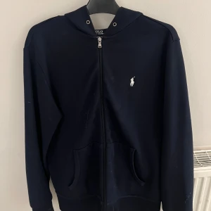 Mörkblå hoodie från Polo Ralph Lauren - Säljer en stilren mörkblå hoodie från Polo Ralph Lauren med dragkedja och det klassiska logotypbroderiet på bröstet. Perfekt för en avslappnad look. Den har långa ärmar och en praktisk huva.