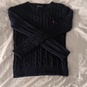 Mörkblå kabelstickad tröja från Polo Ralph Lauren - Säljer en stilren mörkblå kabelstickad tröja från Polo Ralph Lauren. Tröjan har en klassisk design med rund hals och långa ärmar. Den ikoniska loggan i rött finns broderad på bröstet. Perfekt för en elegant och avslappnad look. Tröjan har storlek S och har ordinarie priset 2395 kr men jag säljer den för 500 kr, en perfekt sommar tröja till sena kvällar! Hör av dig vid intresse!