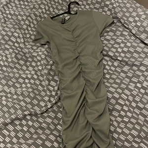 Grön ribbad klänning från H&M - Snygg grön ribbad klänning från H&M med korta ärmar och rynkdetaljer längs framsidan. Perfekt för en stilren look. Klänningen har en figurnära passform och är gjord i ett mjukt material.