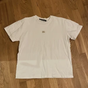 Vit t-shirt från Dolce & Gabbana - Säljer en stilren vit t-shirt från Dolce & Gabbana med ett diskret DG-emblem på bröstet. Perfekt för en minimalistisk look. T-shirten har en klassisk passform och är kortärmad. Storlek S ungefär.