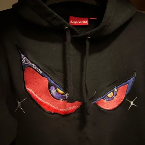 Supreme hoodie - Säljer en svart hoodie från Supreme med ett coolt broderi av ögon i rött och blått på framsidan. Hoodien har en klassisk passform med en stor ficka framtill och justerbar huva. Perfekt för streetwear-entusiaster!
