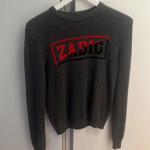 Grå tröja från Zadig & Voltaire - Snygg grå tröja från Zadig & Voltaire med röd och svart text på bröstet. Perfekt för en stilren look. Tröjan har långa ärmar och en rund halsringning.