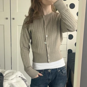 Beige kabelstickad kofta från H&M - Säljer en snygg beige kofta från H&M i storlek XS. Den är ribbad med knappar framtill och har långa ärmar. Perfekt för en stilren look. Tillverkad av återvunnen polyester och bomull. Köpt för 399kr