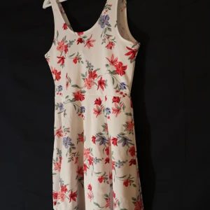 Blommig klänning - Charmig klänning med ett blommigt mönster i rött/rosa, blått och grönt på en vit bakgrund. Perfekt för sommarens varma dagar med sin lätta och luftiga design.