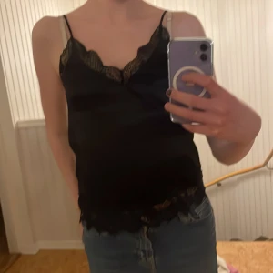 Svart linne med spetsdetaljer - Snyggt svart linne med tunna axelband och vacker spets längs halsringningen och nederkanten. Perfekt för en stilren look.