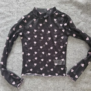 Svart topp med hjärtmönster från Minga London  - Svart, genomskinlig långärmad topp med rosa hjärtan. Den har en hög krage och en vågig kant. Storlek S. Sparsamt använd, mycket fint skick.