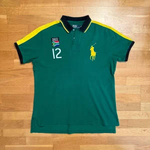 South Africa  Polo Ralph Lauren - Säljer en grön pikétröja från Polo Ralph Lauren med gula detaljer och broderad logga. Tröjan har korta ärmar och nummer 12 tryckt på både fram- och baksidan. Perfekt för en sportig look! Skick 8.5/10 finns inga fel 