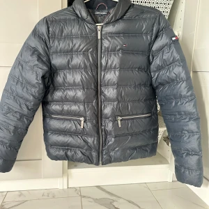 Mörkblå dunjacka från Tommy Hilfiger - Snygg mörkblå dunjacka från Tommy Hilfiger med dragkedja och två fickor framtill. Insidan har ett coolt mönster med blixtar, hjärtan och stjärnor i olika färger. Perfekt för kyliga dagar när du vill hålla stilen. 🧥✨