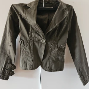 Olivgrön kavaj med knappar - Snygg olivgrön kavaj med knappar framtill och på ärmarna. Den har en klassisk krage och två fickor med knappdetaljer. Perfekt för en stilren look.