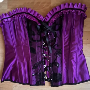 Fin lila korsett! - Jättefin lila korsett i satin och svart spets, mjuka skenor fram med hakar och snörning bak som kan justeras flera dm. Mått: H: 40 cm B: 80 cm runtom på innersta snörning med utrymme för extra 25 cm. Perfekt skick!